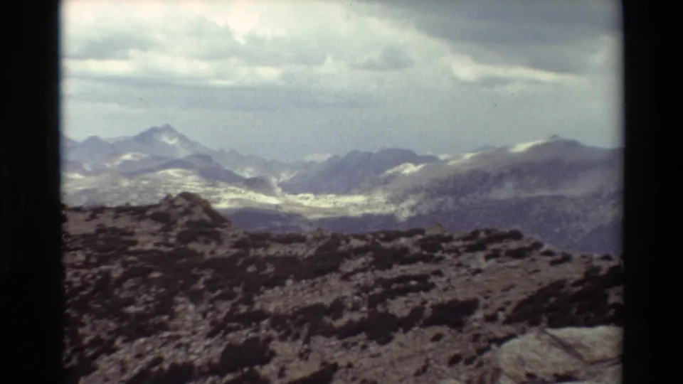 Vintage 8mm view across a rocky overlook toward snow-marked mountains, with dark clouds and soft film color.