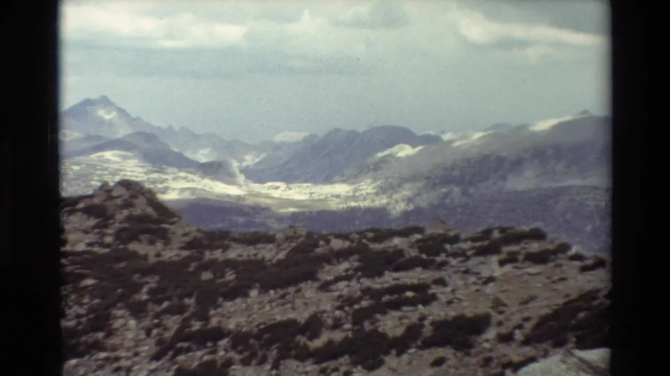 Home-movie mountain landscape frame with a closer view of snowy ridges, muted sky, and rough alpine ground in front.