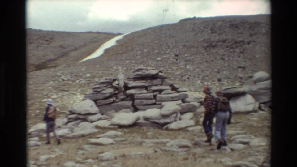 Three bundled hikers with backpacks move across barren rocky alpine ground near a small stone cairn and a narrow snow patch.