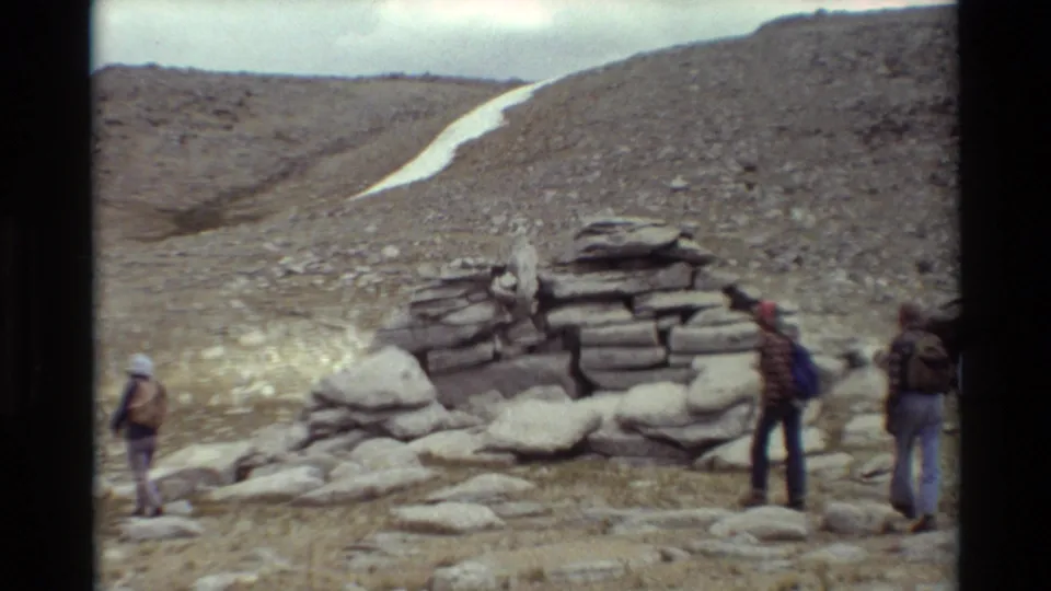 Three distant hikers in outdoor clothing and backpacks stand near a stone cairn on rocky alpine terrain, with a narrow snow patch behind them.