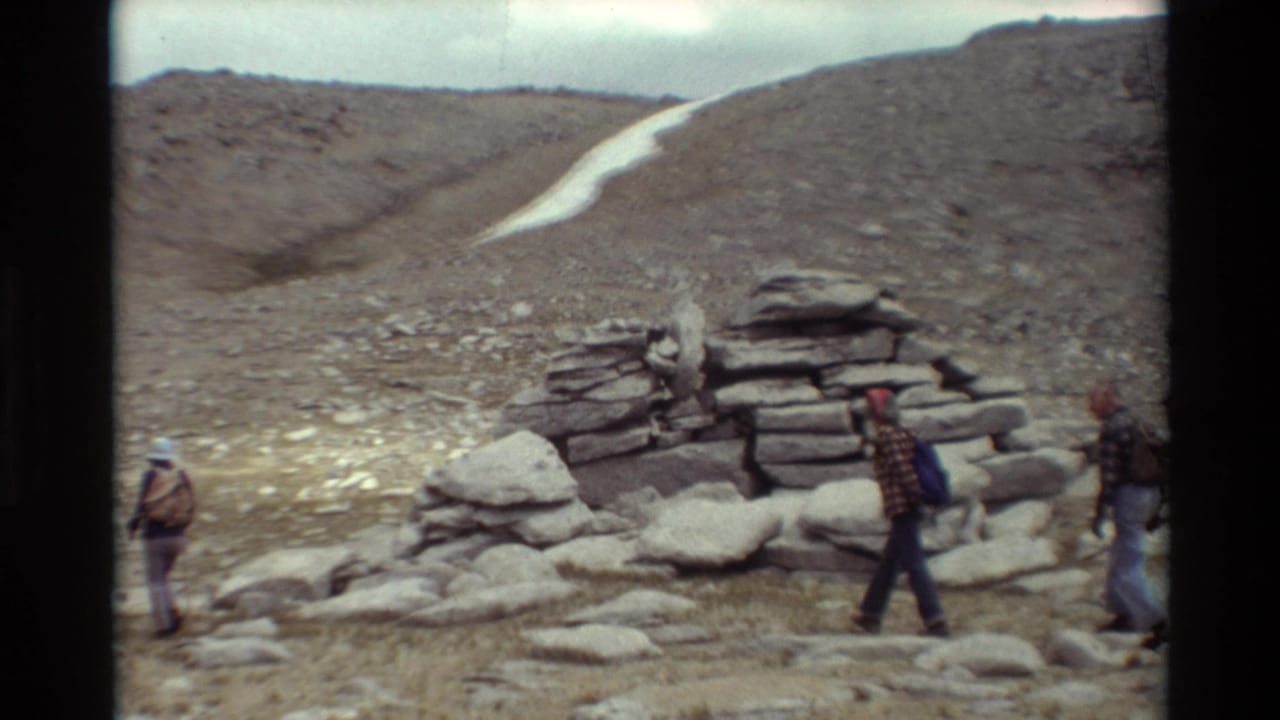 1981 8mm home-movie frame of hikers with backpacks passing a stone cairn in a rocky mountain landscape.