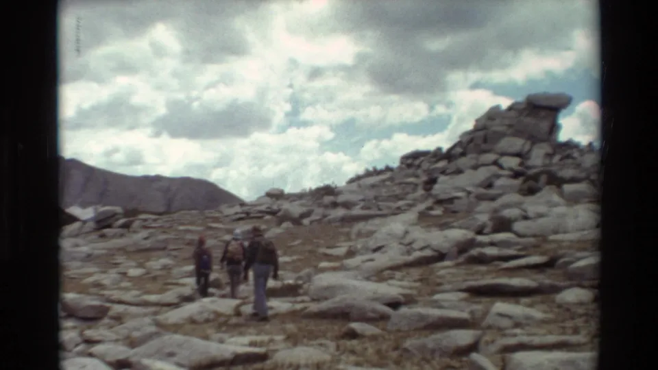 A small group of hikers crosses a rocky mountain area toward a large boulder outcrop in 1981 8mm footage.