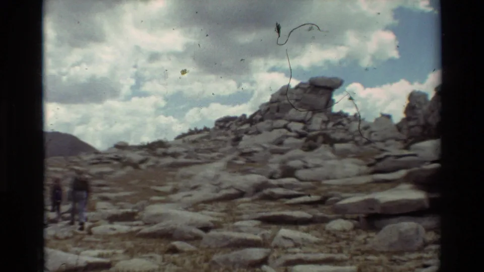 Hikers walk away through a rocky alpine-looking landscape with a prominent stone outcrop and cloudy sky.
