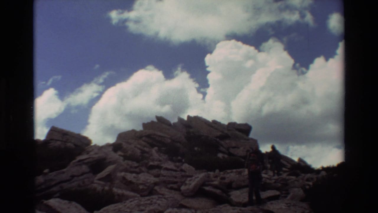 1981 8mm home-movie frame of hikers moving down dark rocks under a deep blue sky with billowing clouds.