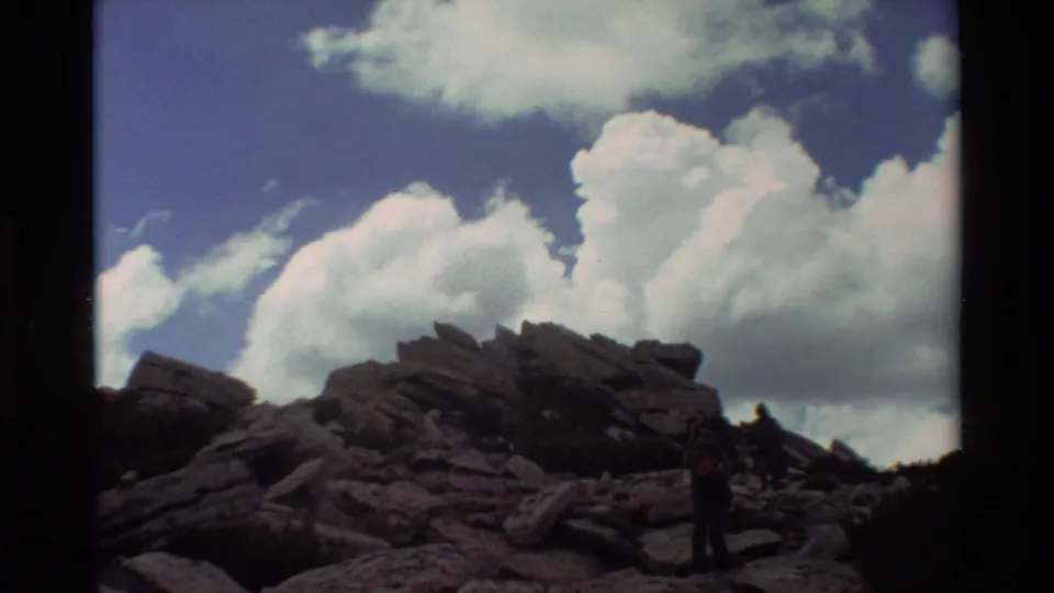1981 8mm home-movie frame of hikers moving down dark rocks under a deep blue sky with billowing clouds.