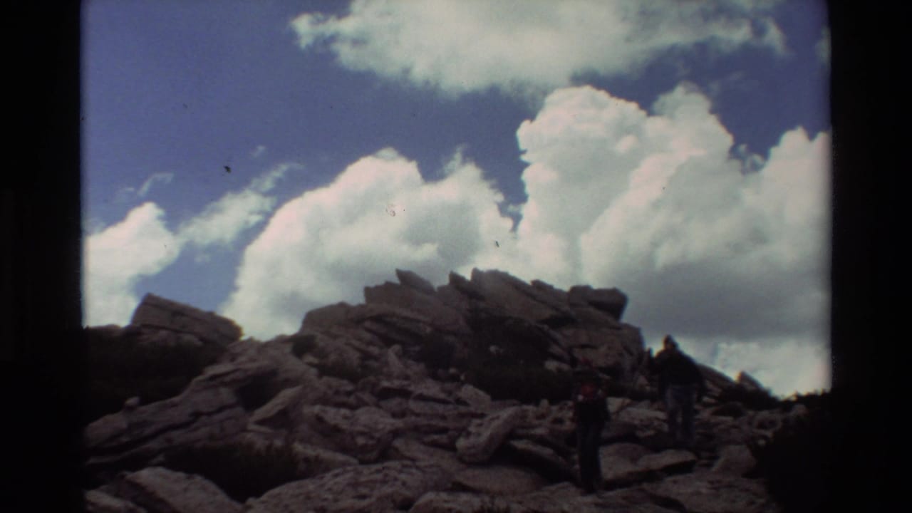 Two hikers move across rocks under a bright cloudy sky.