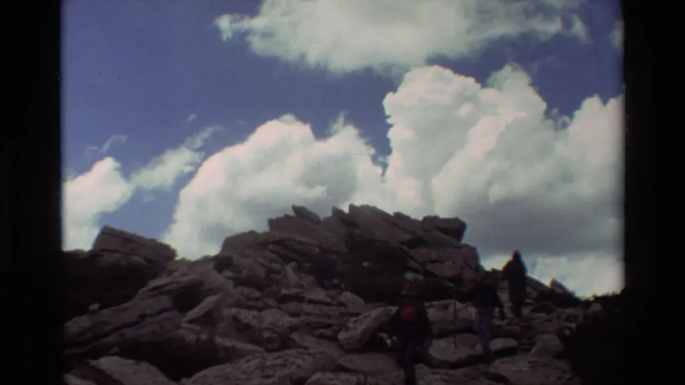 Several distant hikers appear on a rocky slope in a 1981 8mm outdoor recreation scene with large clouds behind them.