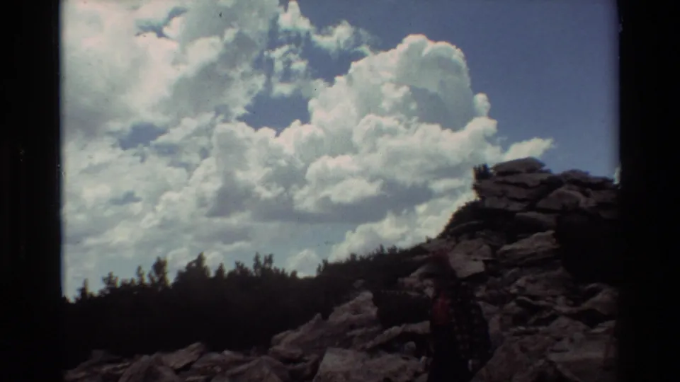 A distant hiker stands beside a rocky rise, with dark trees and billowing clouds visible in the 1981 home-movie frame.