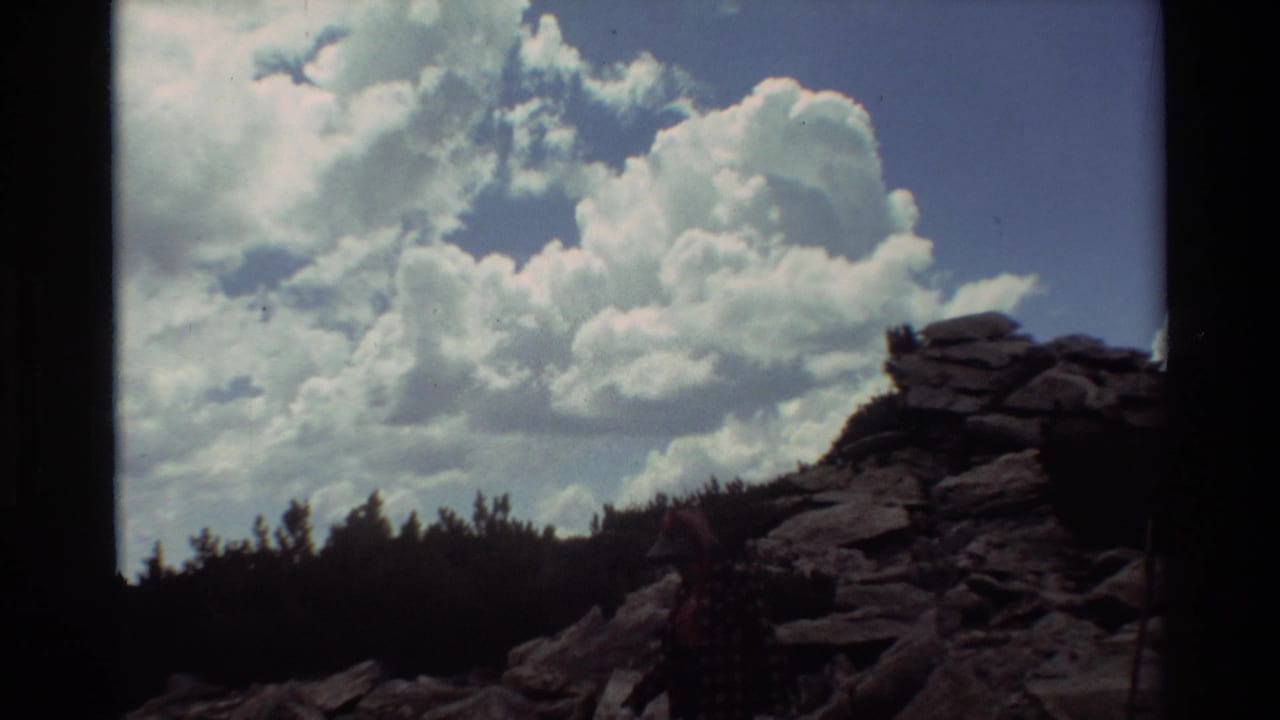 1981 8mm home-movie frame of a rocky mountain trail with a partly visible hiker, treeline, blue sky, and large cumulus clouds.