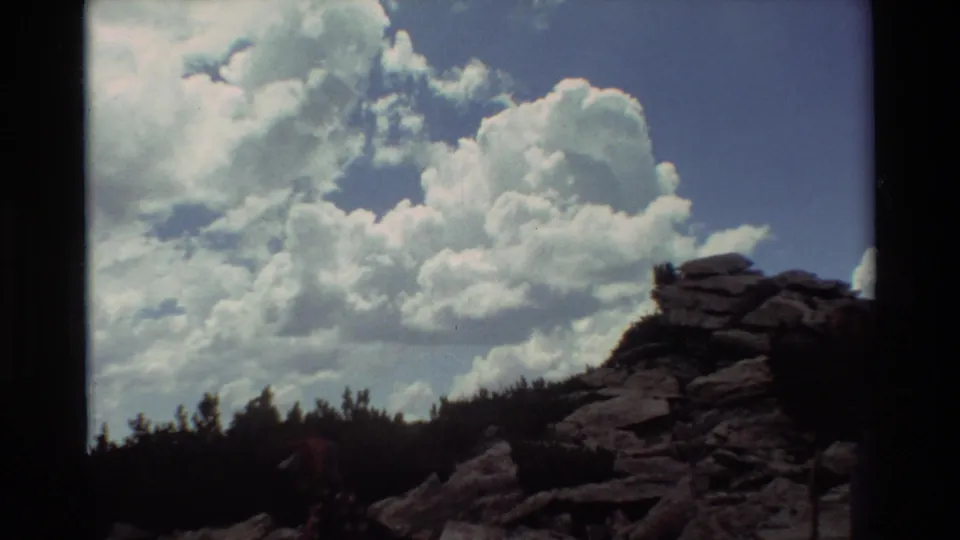1981 8mm home-movie frame showing rugged rocks, a dark treeline, bright cloud formations, and a hiker partially obscured near the trail.