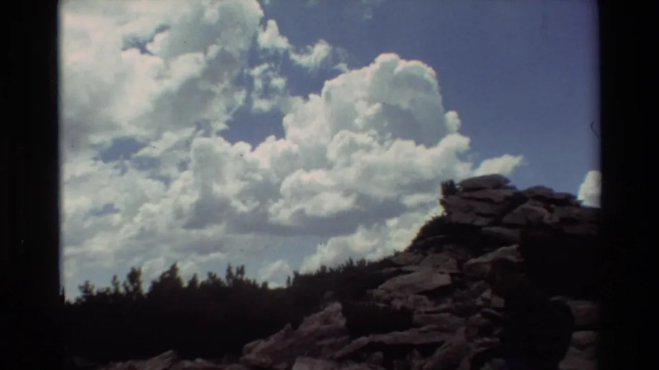 1981 8mm home-movie frame of a hiker on a rocky mountain trail below a treeline and broad white clouds.