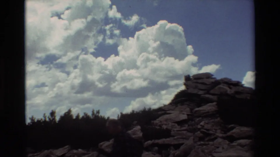 1981 8mm home-movie frame showing two hikers on uneven rocks, backed by a dark treeline and large white clouds.