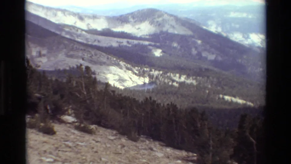 1981 8mm home-movie view across a dry slope and conifer valley toward snow-patched mountain ridges in the listed British Columbia setting.