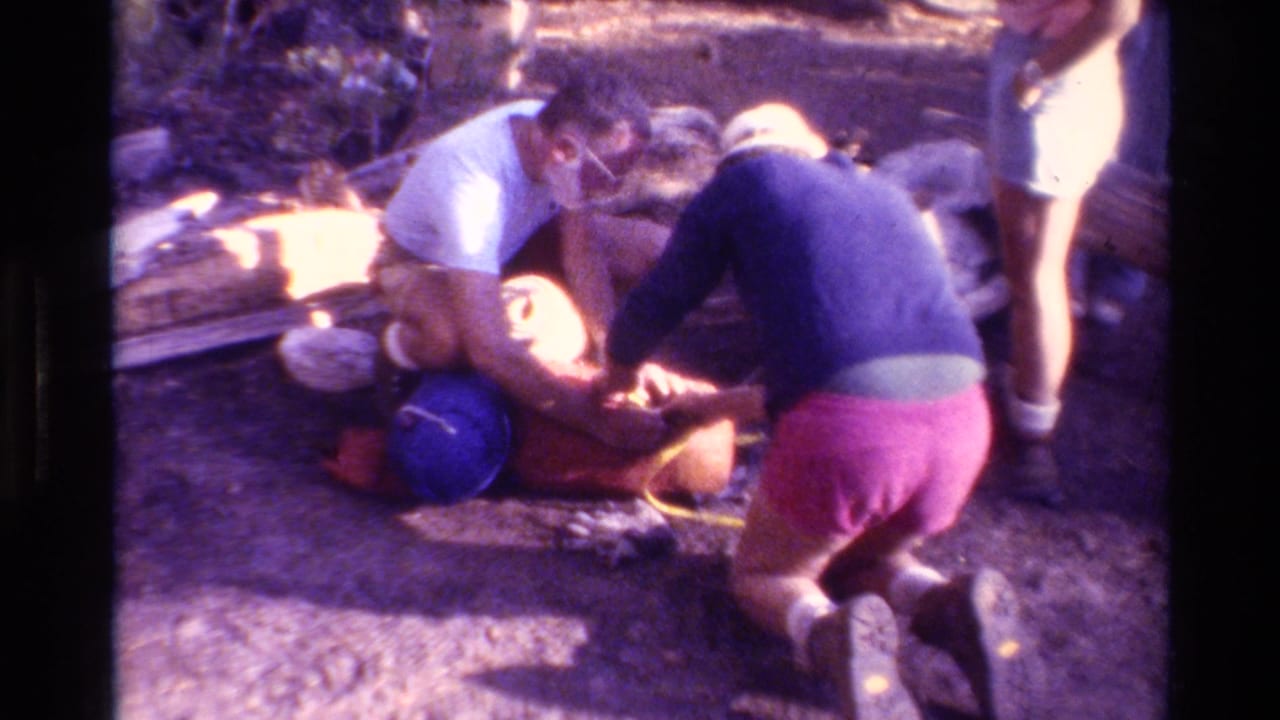 1981 home-movie frame of hikers in a wooded British Columbia setting kneeling beside packs and bundled camping gear.