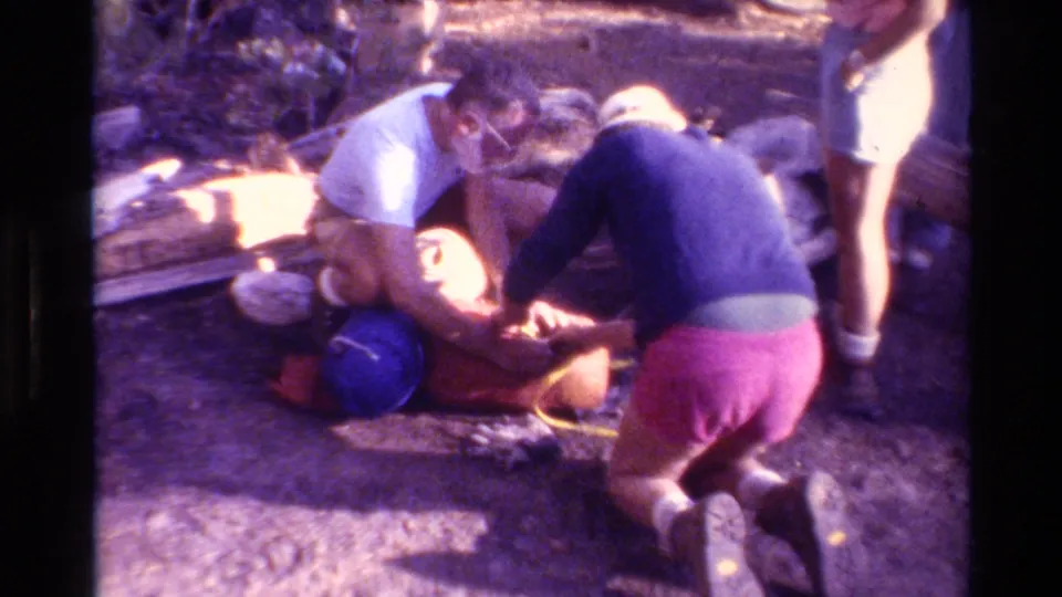 1981 home-movie frame of hikers in a wooded British Columbia setting kneeling beside packs and bundled camping gear.