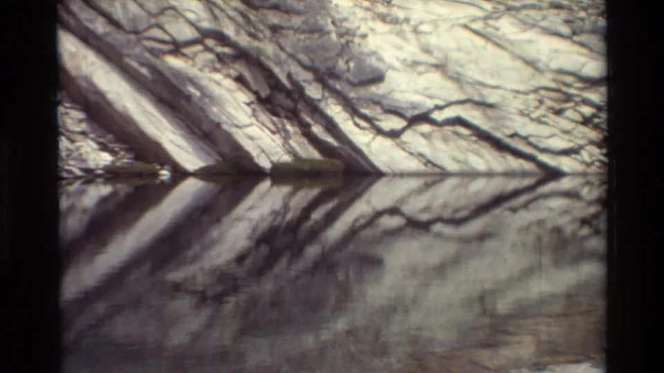 1981 8mm frame listed as British Columbia, with layered rock forms and bare branch reflections mirrored in the stream surface.