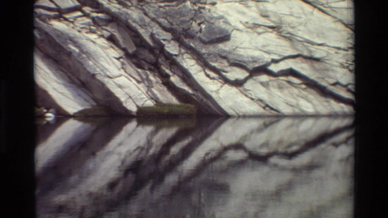 Waterside cliff detail from the supplied 1981 8mm clip, showing layered gray rock reflected in still water.
