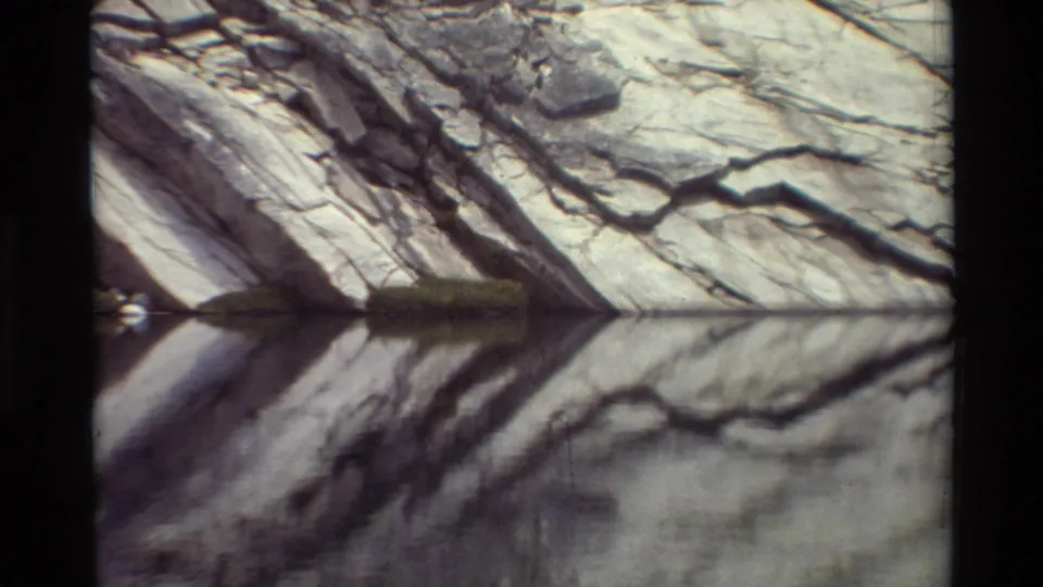 Waterside cliff detail from the supplied 1981 8mm clip, showing layered gray rock reflected in still water.
