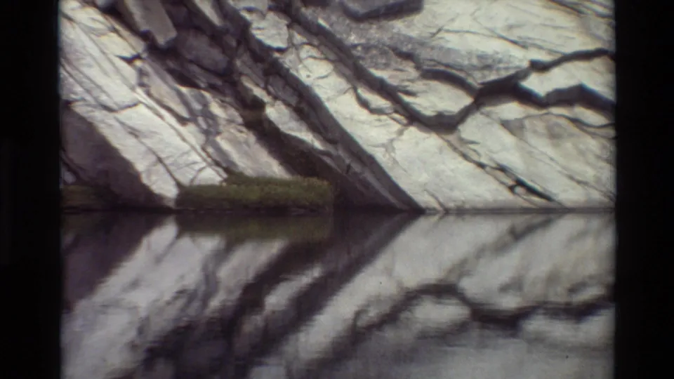 Frame from a listed British Columbia 8mm landscape clip with angled rock, still water, and a small green patch at the cliff base.