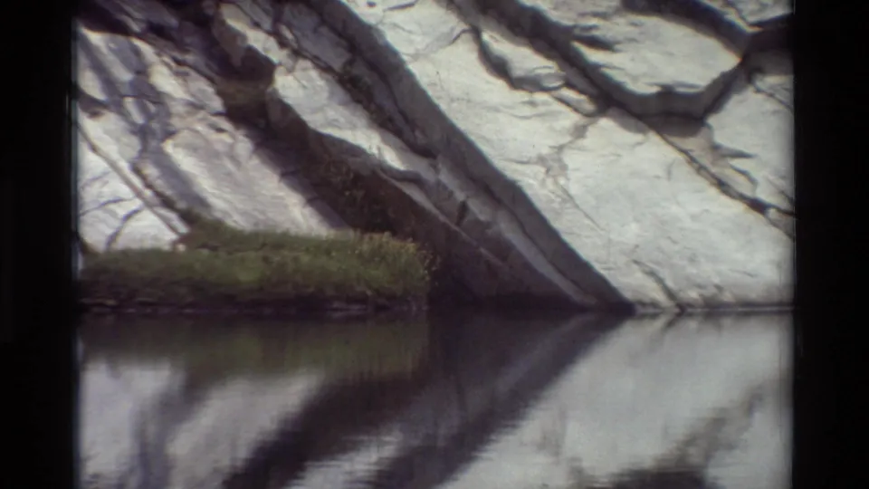 Closing 8mm frame showing the cliff base, a patch of green vegetation, and the rock face reflected in calm water.