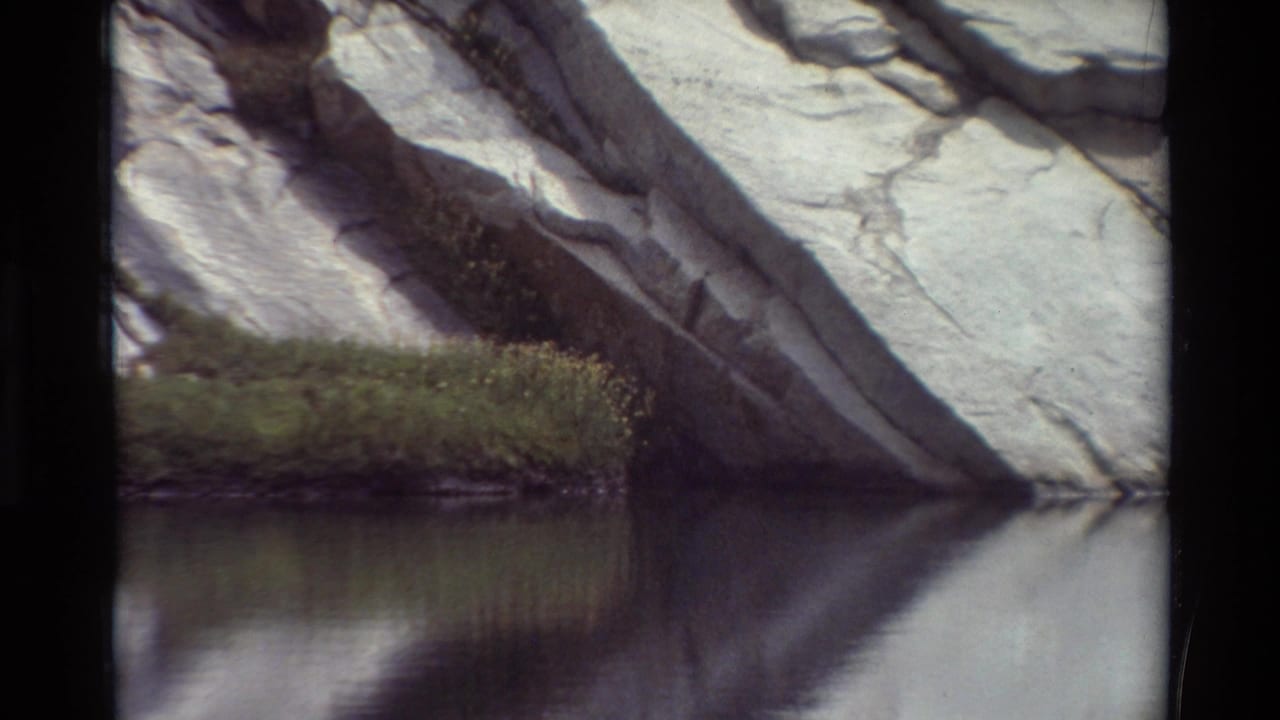 1981 8mm frame listed as British Columbia showing pale rock above still water and a small grass-covered edge with flowers.