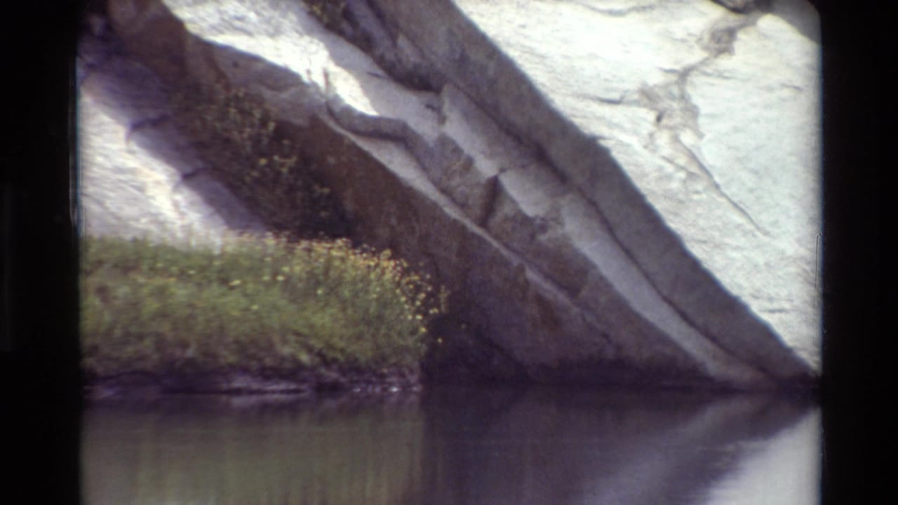 Vintage 8mm view of an angled rock outcrop over calm water with grass and small yellow flowers along the edge.