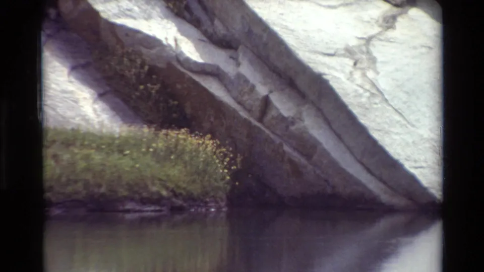 Vintage 8mm view of an angled rock outcrop over calm water with grass and small yellow flowers along the edge.