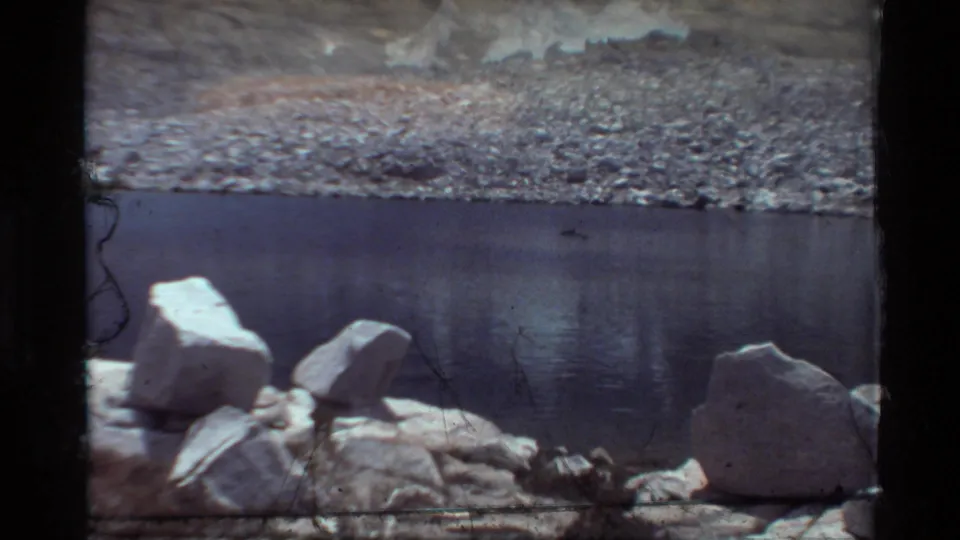 Wider 1981 8mm view of a rocky lakeshore or pond edge with still dark water and pale stones in the foreground.