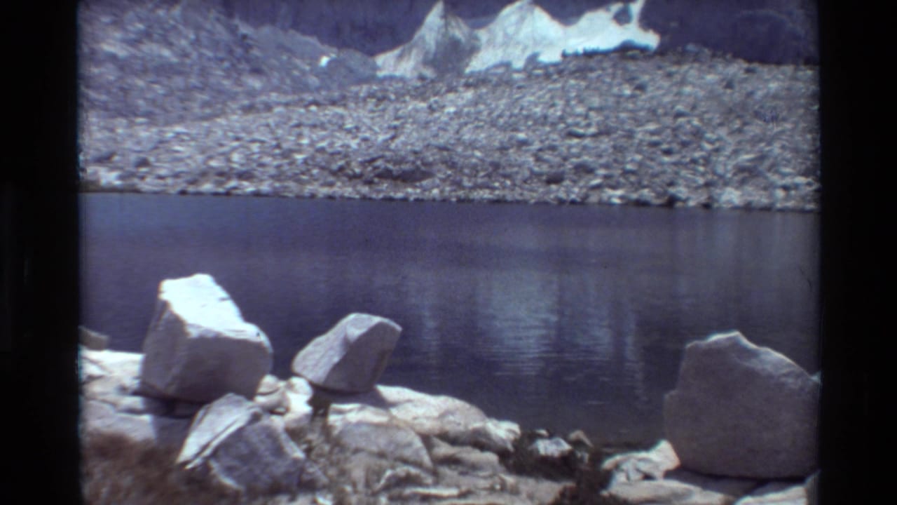 1981 8mm home-movie view of an alpine lake in the stated British Columbia region, with boulders, rocky shoreline, snow patches, and mountains.