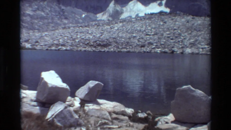 1981 8mm home-movie view of an alpine lake in the stated British Columbia region, with boulders, rocky shoreline, snow patches, and mountains.