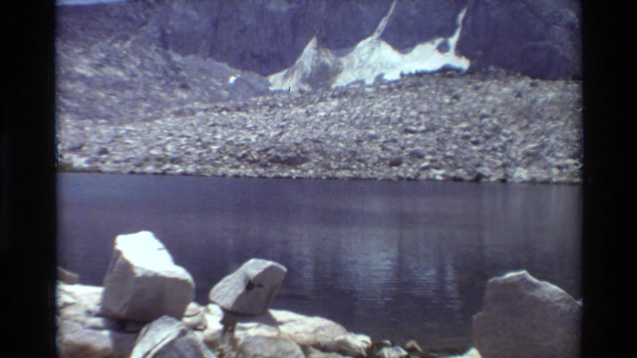 8mm frame of a quiet alpine lake with bright foreground rocks, a stony far shore, and snow against steep mountain terrain.