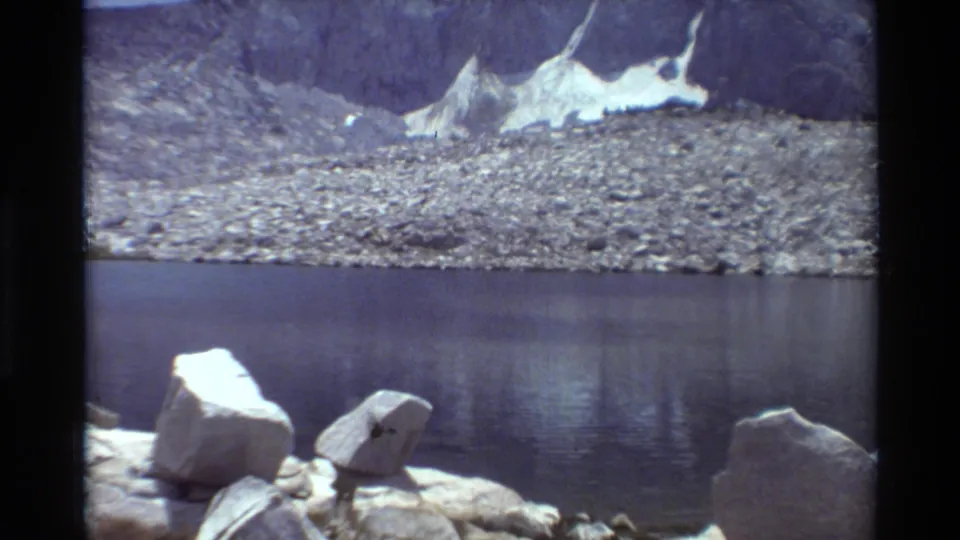 8mm frame of a quiet alpine lake with bright foreground rocks, a stony far shore, and snow against steep mountain terrain.