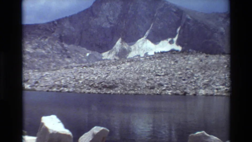 Vintage 8mm alpine lake view, showing dark water, a broad rocky slope, jagged mountains, and remnant snow under cloudy light.
