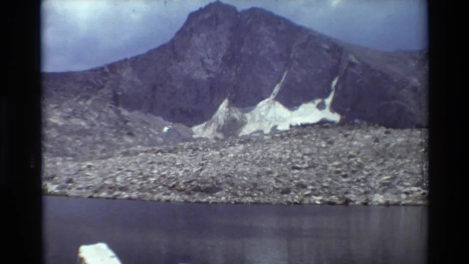 1981 8mm home-movie frame of an alpine lake and rugged mountain slope with visible snow patches and muted cloudy light.