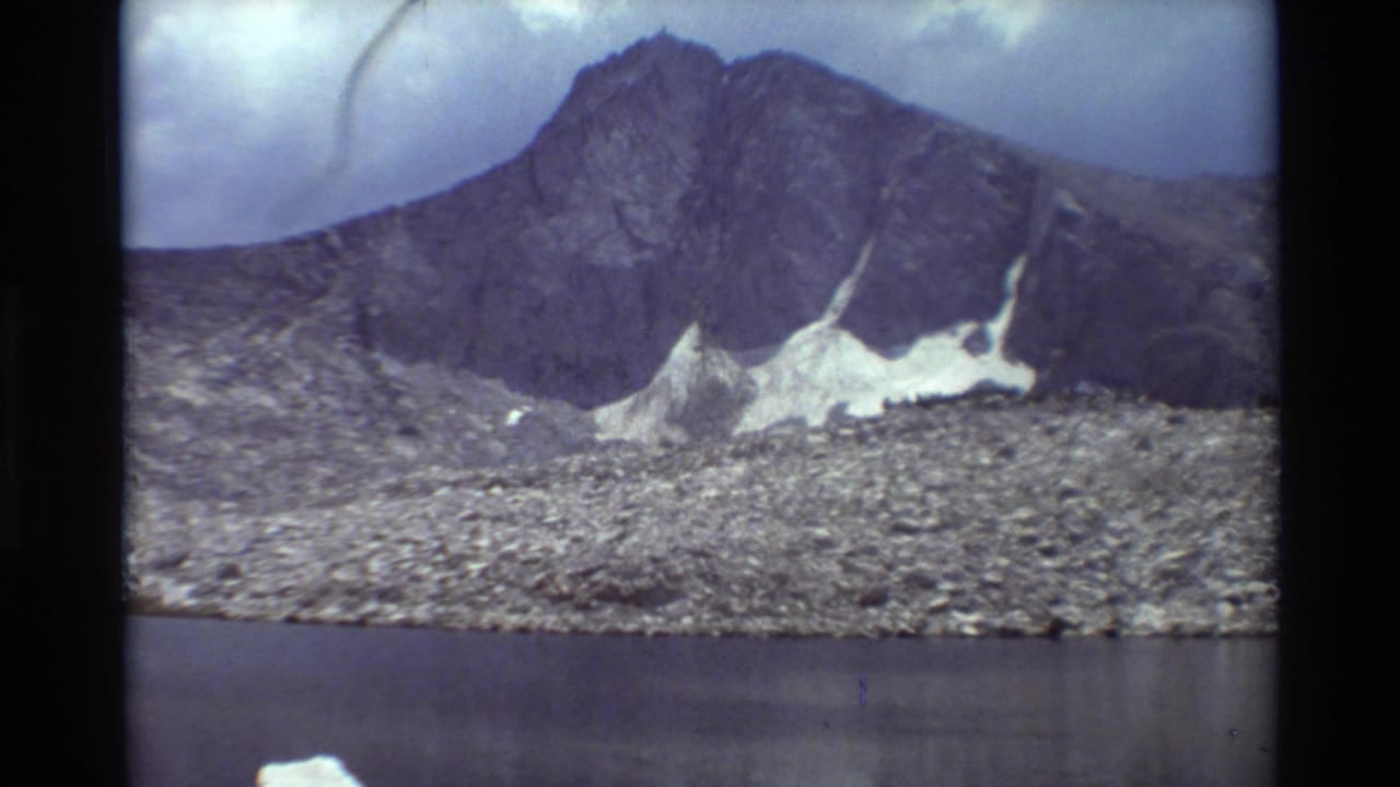 Cataloged 1981 8mm frame showing an alpine mountain, snow patches, rocky shore, and calm lake water in British Columbia footage.