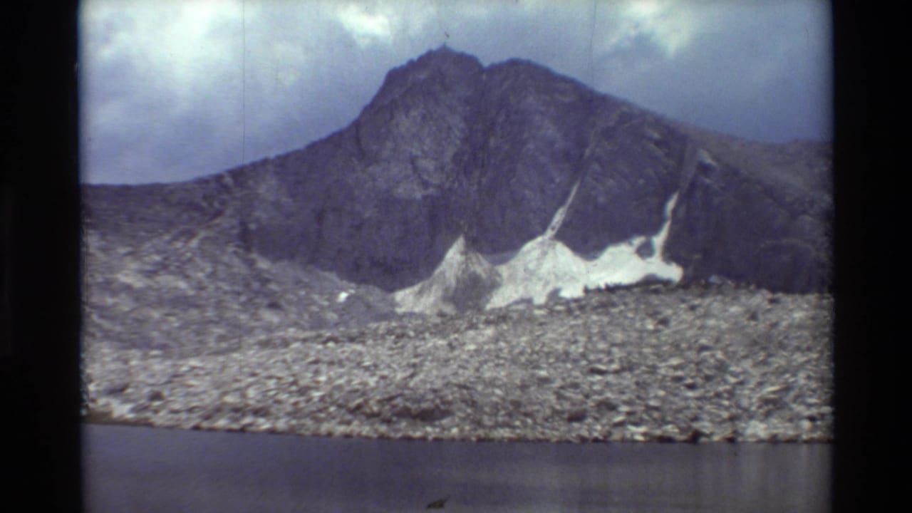 Cataloged 1981 8mm view of a dark rocky ridge, bright snow remnants, and still water beneath a clouded sky.
