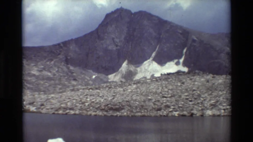 Cataloged British Columbia 8mm frame with a rocky mountain, pale snow patches, shoreline stones, and dark lake water.