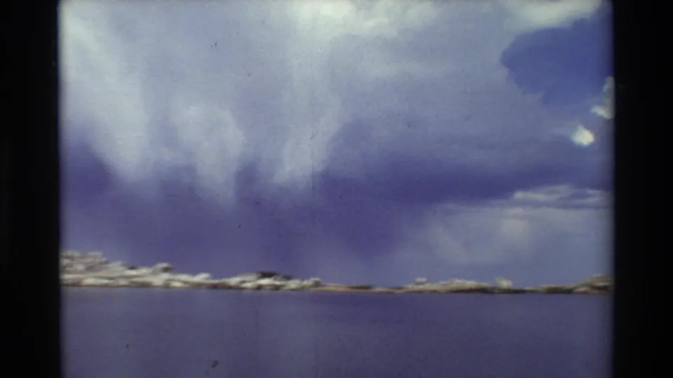 Cataloged 1981 8mm frame showing heavy clouds over open water and a low rocky shoreline in a scenic lake setting.