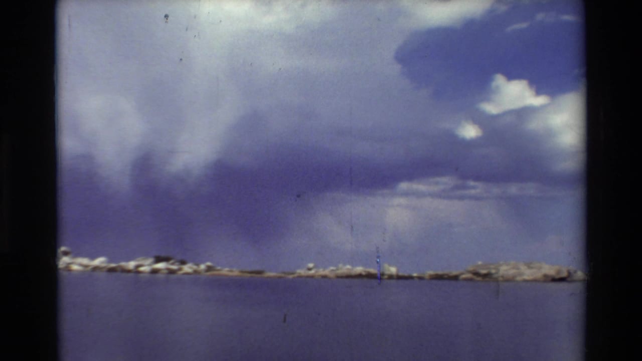 1981 8mm home-movie view of a calm mountain lake, distant rocky shore, and dramatic cloud formations overhead.