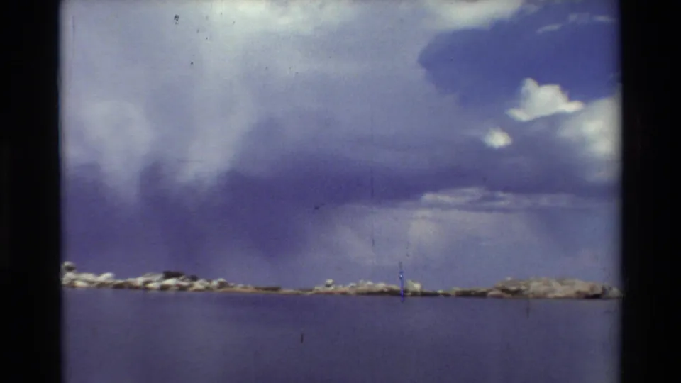 1981 8mm home-movie view of a calm mountain lake, distant rocky shore, and dramatic cloud formations overhead.