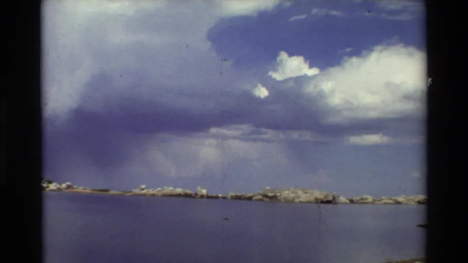 1981 8mm landscape frame showing a still lake, rocky far shore, and layered cumulus clouds above the water.