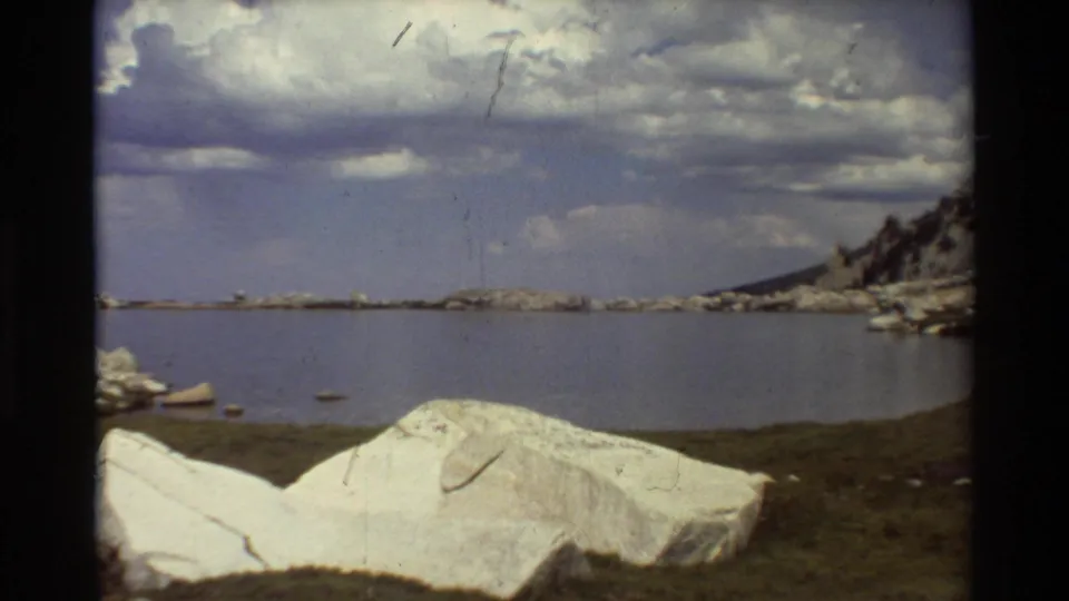 1981 8mm home-movie frame with pale boulders in the foreground, calm lake beyond, and clouds over a rugged shore.
