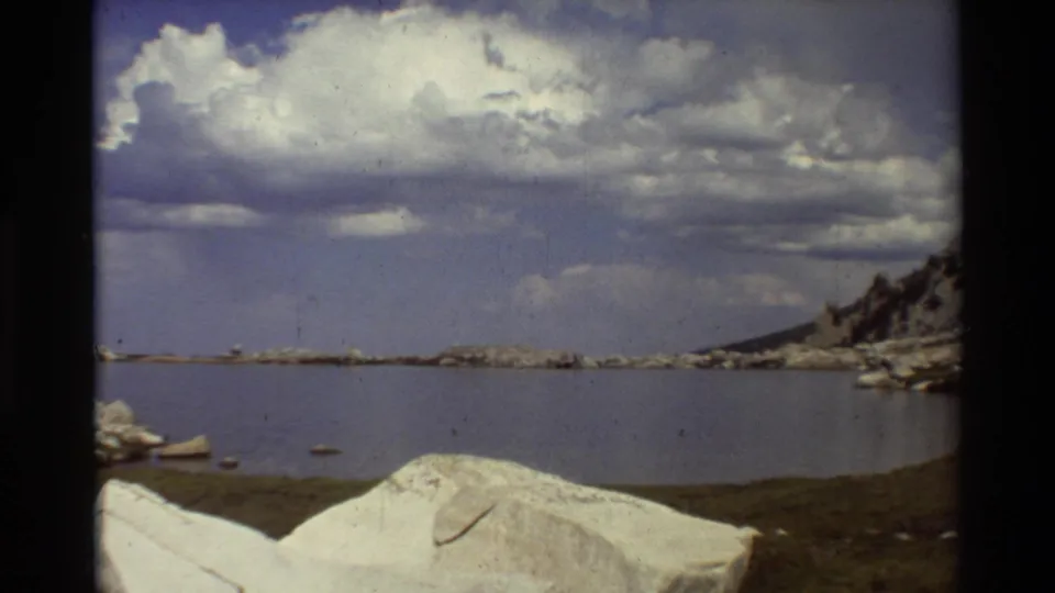 1981 8mm lakeshore view in British Columbia, with foreground boulders, still water, distant rocky land, and cumulus clouds.