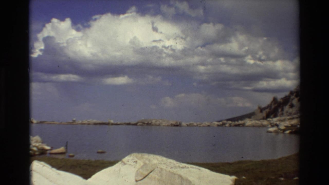 1981 8mm frame of a calm mountain lake with pale boulders, cloud-filled sky, and a rocky shoreline in the supplied British Columbia location metadata.