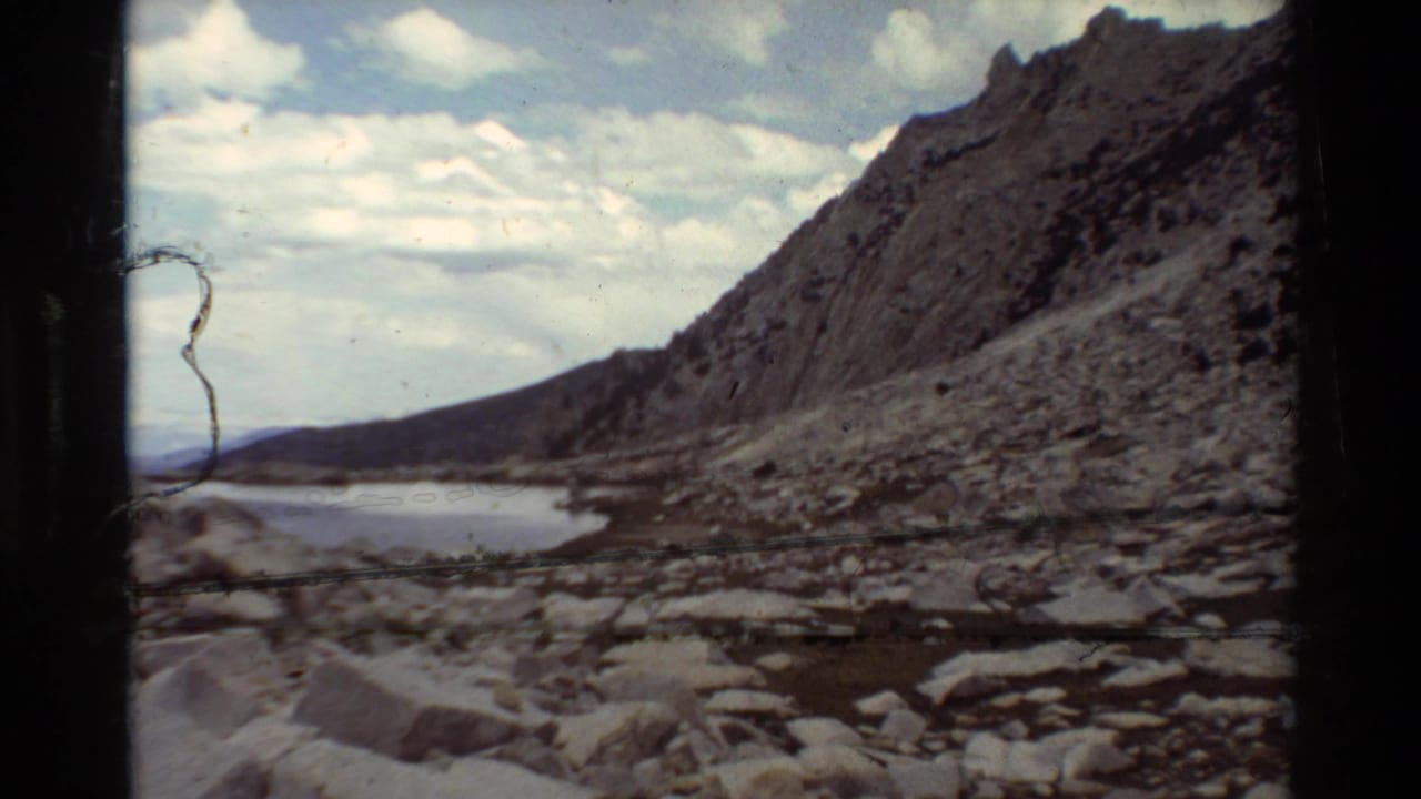 Rocky lakeshore and steep sparsely vegetated slope seen in a 1981 8mm home-movie frame, with water curving along the base of the terrain.