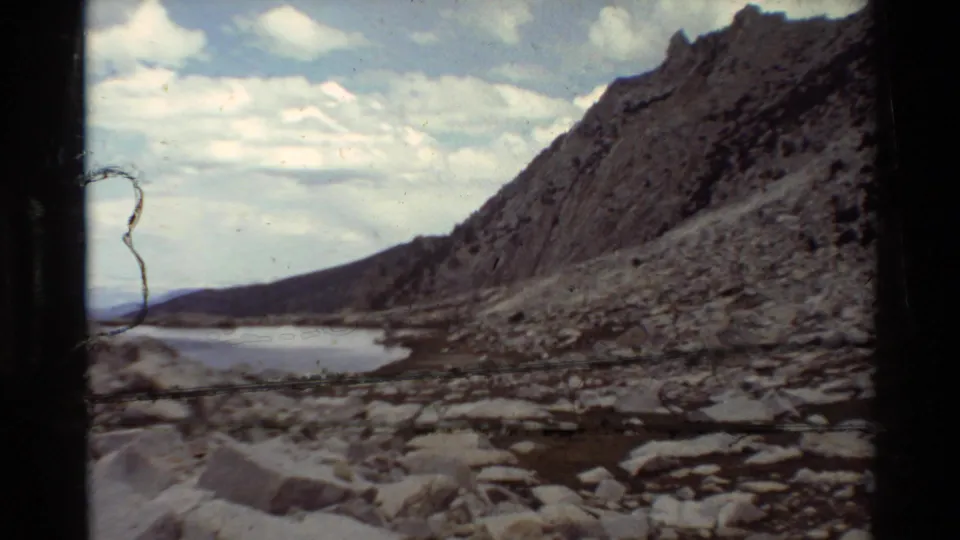 Rocky lakeshore and steep sparsely vegetated slope seen in a 1981 8mm home-movie frame, with water curving along the base of the terrain.