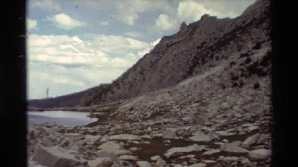 1981 8mm frame looking along a rocky lakeshore toward a steep, mostly bare slope with water at the left edge and clouds overhead.