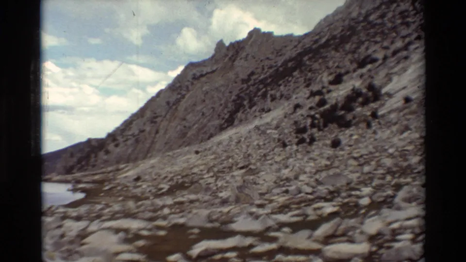 Final 1981 8mm frame of a rugged boulder field and steep rocky mountainside beside the lake, with pale clouds and visible film grain.