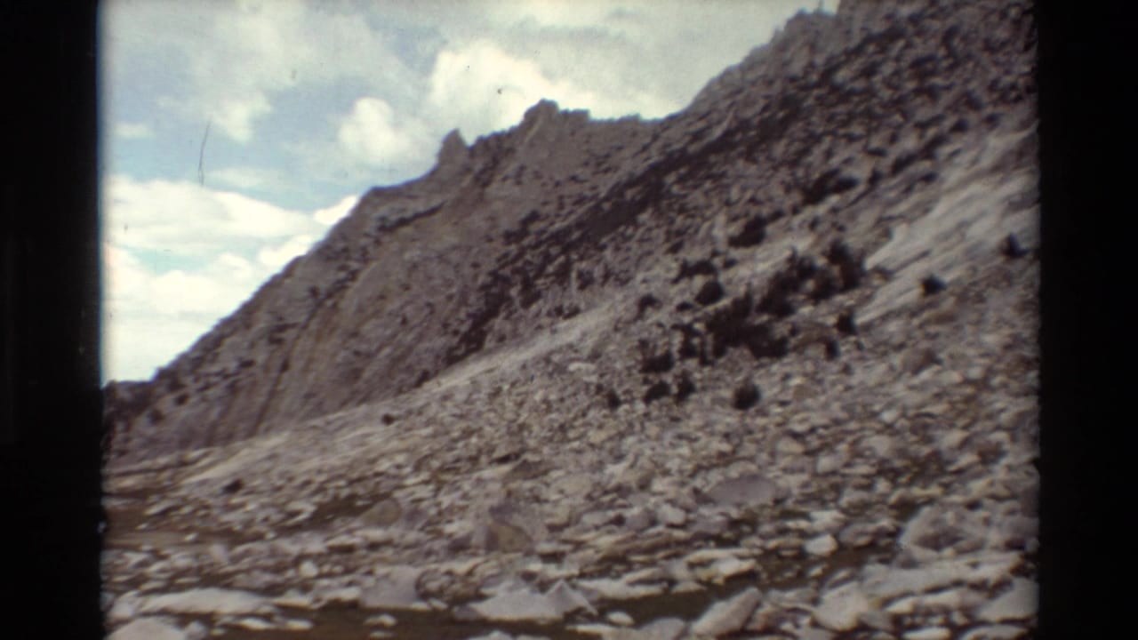 1981 8mm home-movie view of a rocky mountain slope in the listed British Columbia setting, with a pale trail and cloudy sky.