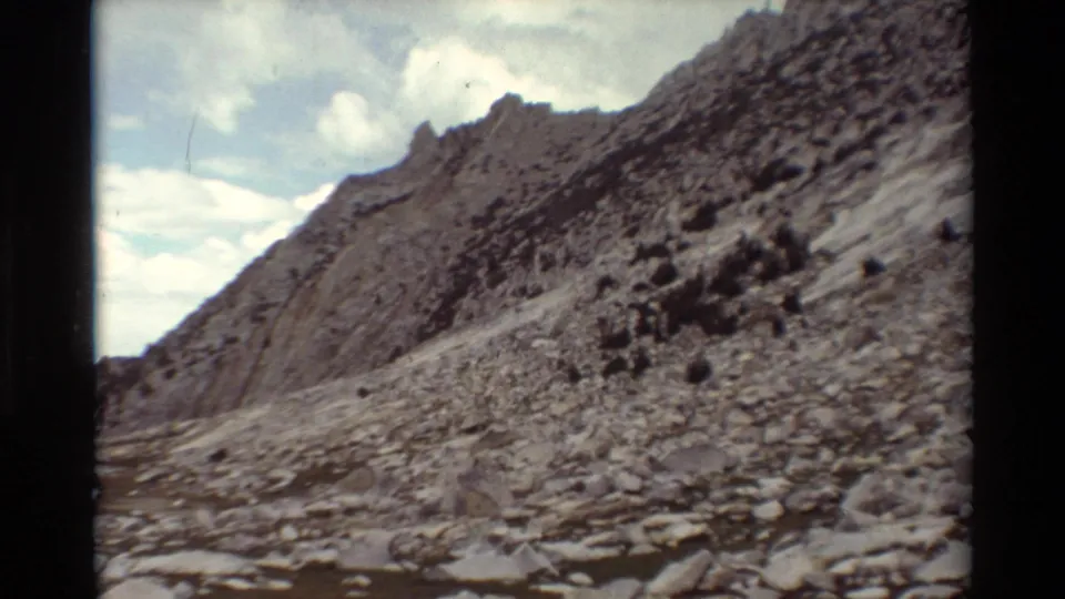 1981 8mm home-movie view of a rocky mountain slope in the listed British Columbia setting, with a pale trail and cloudy sky.
